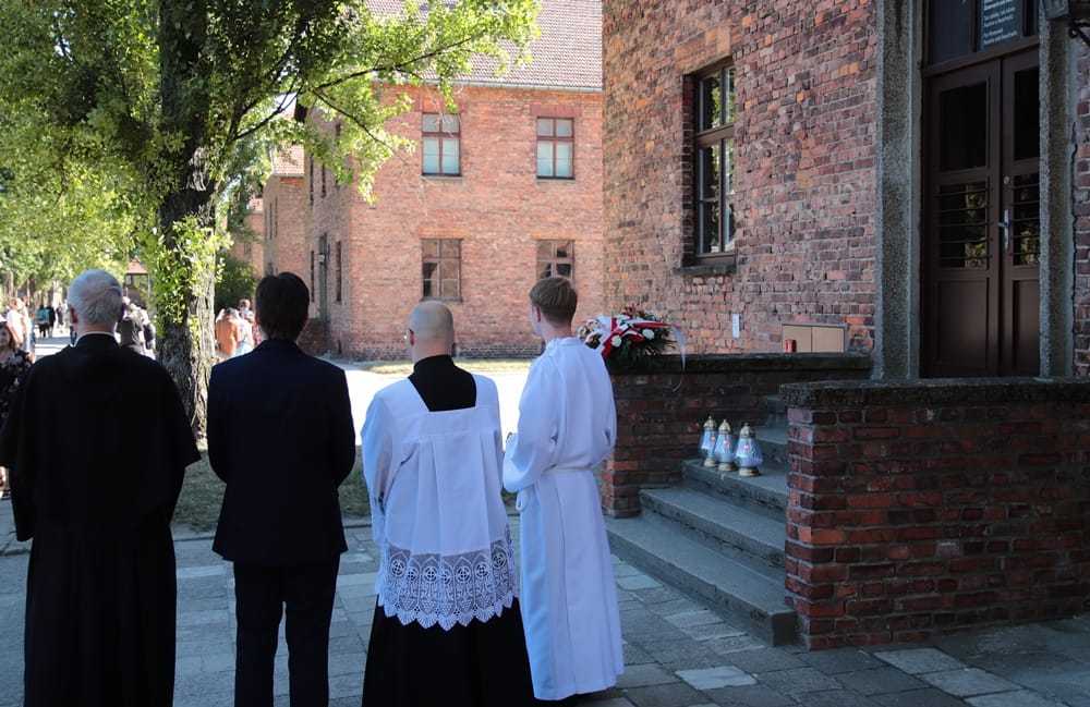 Rückenansicht von vier Personen – einem Ordensbruder in schwarzer Kutte, einem Mann im Anzug sowie zwei Ministranten in weiße Gewänder gekleidet – vor der Treppe und dem Eingang eines historischen Backsteingebäudes (Block 14) in Auschwitz.