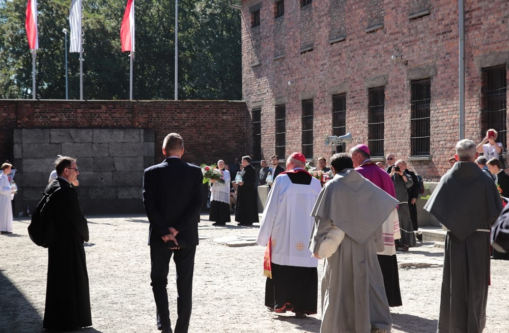 Der Innenhof aus Kies zwischen zwei Backsteinbauten (Block 10 und 11) mit der grau rekonstruierten Todeswand im Hintergrund. Mehrere Geistliche in liturgischen Gewändern, Ordensbrüder und Würdenträger stehen im Hof versammelt. Im Hintergrund wehen die polnischen Flaggen.