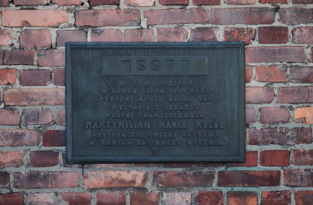 Eine dunkle Metall-Gedenktafel an einer roten Backsteinwand des ehemaligen Blocks 14a in Auschwitz. Die Inschrift auf Polnisch erinnert an die Häftlingsnummer 16670 von Maximilian Kolbe und seinen freiwilligen Schritt in den Tod für einen Mithäftling.
