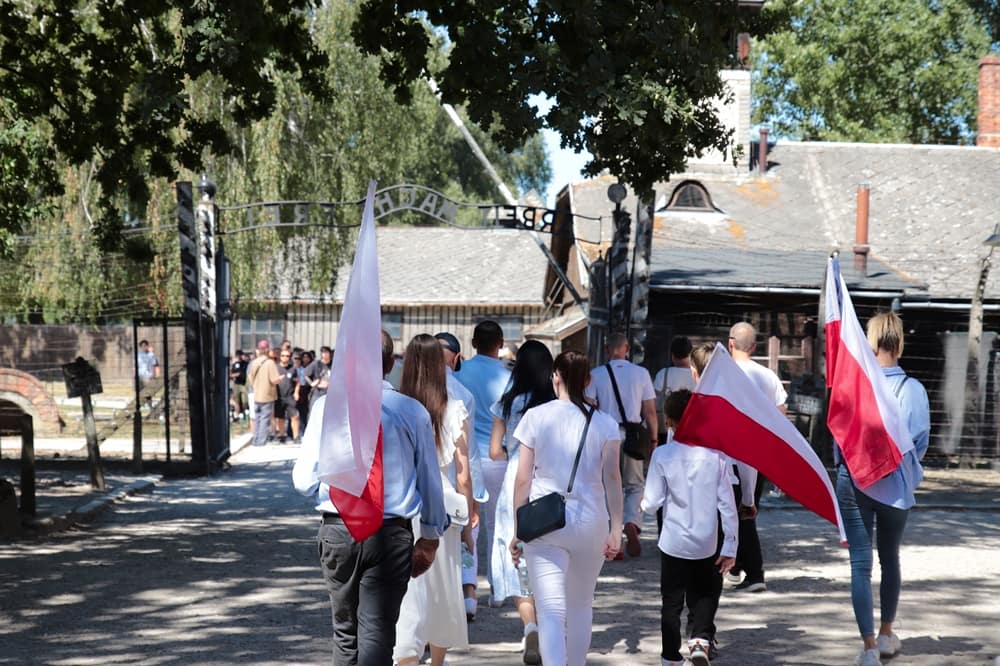 Blick von innen auf das eiserne Haupttor des Stammlagers Auschwitz I mit dem Schriftzug „Arbeit macht frei“ im Bogen. Eine Gruppe von Besuchern und Teilnehmern mit polnischen Flaggen geht durch das Tor nach draußen.