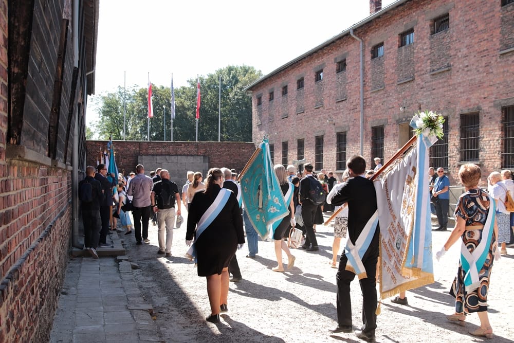 Eine Gruppe von Menschen zieht in einer Prozession durch den Hof vor der Backsteinfassade von Block 11. Mehrere Personen tragen blau-weiße Schärpen und halten große Prozessionsfahnen mit religiösen und Gedenk-Motiven.